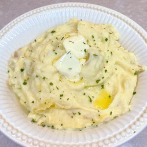 white bowl containing mashed potatoes with two pats of butter melting on top and sprinkled with chopped parsley