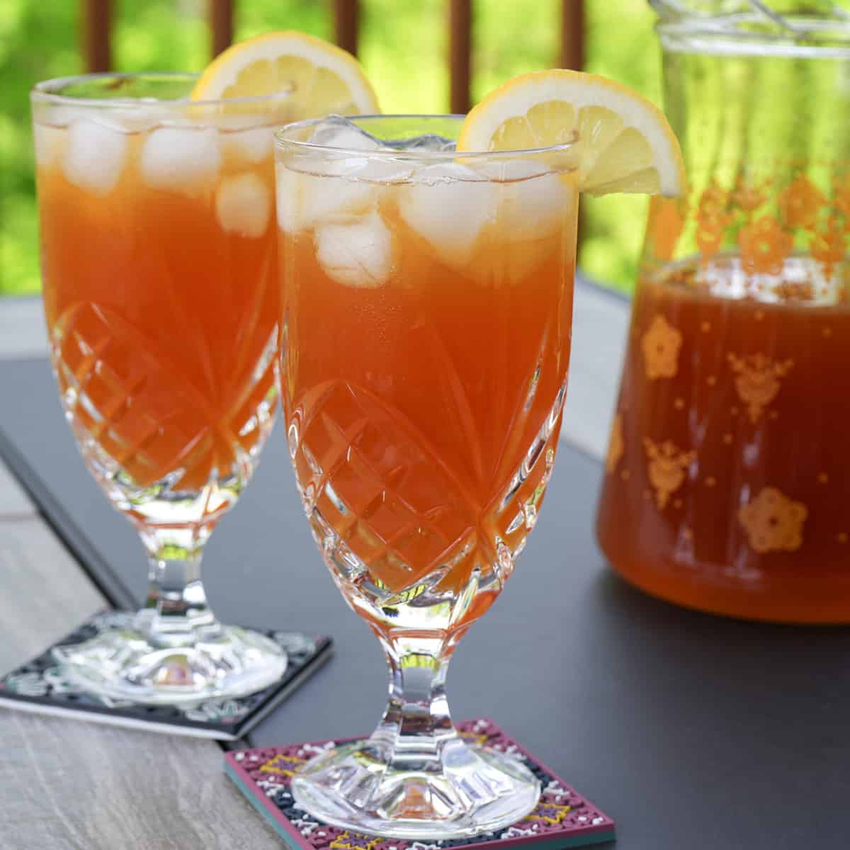 two crystal glasses of lemonade iced tea with lemon garnish and tea pitcher in background.